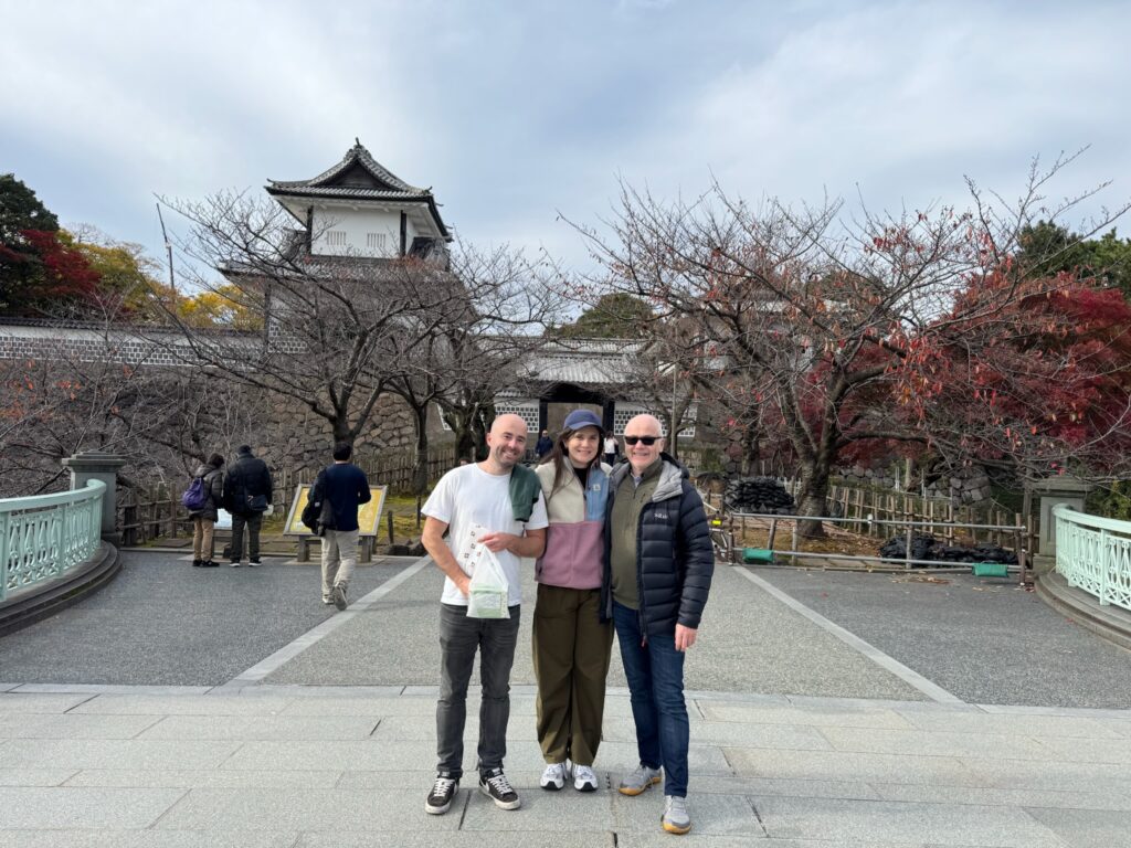 Three travelers standing in front of Kanazawa Castle, smiling with autumn trees in the background during a walking tour in Kanazawa.